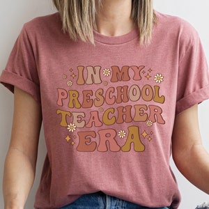 May include: A pink t-shirt with the text "In My Preschool Teacher Era" in a retro font with flowers and stars.