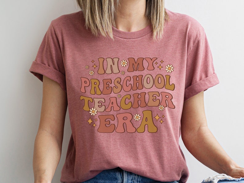 May include: A pink t-shirt with the text "In My Preschool Teacher Era" in a retro font with flowers and stars.
