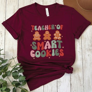 May include: Burgundy t-shirt with a graphic design that says "Teacher of Smart Cookies" with gingerbread men illustrations.