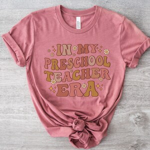 May include: A pink t-shirt with the text "In My Preschool Teacher Era" in a retro font with brown, orange, and yellow colors. The text is surrounded by small white flowers and gold stars.