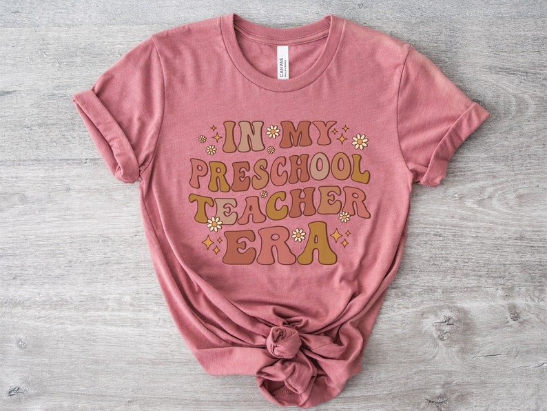 May include: A pink t-shirt with the text "In My Preschool Teacher Era" in a retro font with brown, orange, and yellow colors. The text is surrounded by small white flowers and gold stars.
