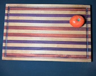 May include: A wooden cutting board with a striped pattern in shades of brown, purple, and light wood. A single red tomato sits on the board.