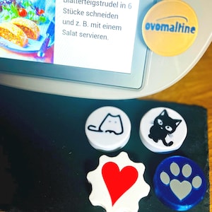 Op de afbeelding: Een verzameling decoratieve magneten met verschillende ontwerpen. Eén magneet toont de tekst "ovomaltine" op een gele achtergrond. Andere magneten tonen een kat, een hart en een pootafdruk. De magneten liggen op een zwart oppervlak bij een digitaal scherm.