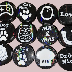 Peut inclure: Une collection de boutons noirs et circulaires avec divers motifs et textes blancs. Les motifs comprennent une manette de jeu, des empreintes de pattes avec un cœur, un pingouin, le mot "Love", un fer à cheval, une tête de cheval, un hibou, un chapeau de chef et les mots "Mr & Mrs", "Dog Mom", "Cat Mom", et "Drück Mich".