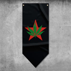 Puede incluir: Bandera negra con una estrella roja y un diseño de hoja de cannabis verde. La bandera tiene una base puntiaguda y dos ojales en la parte superior para colgar. El fondo es una pared gris texturizada.