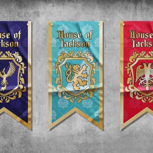Op de afbeelding: Drie banners met de tekst "House of Jackson" in gouden letters op een blauwe, turquoise en rode achtergrond. Elke banner heeft een ander gouden wapenschild met een feniks, een leeuw en een adelaar.