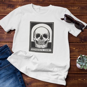 May include: White t-shirt featuring a black and white illustration of a skull with the text "Memento Mori" underneath.