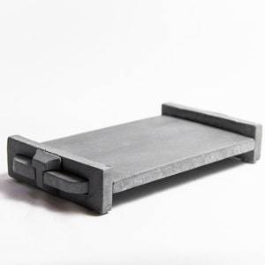 May include: A grey concrete tray with a rectangular shape, raised edges, and rounded handles. The minimalist design is suitable for serving or display. The tray is presented against a white backdrop.