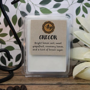 May include: A white wax melt in a clear plastic container with a label that reads "GREGOR Bright lemon zest, sweet grapefruit, rosemary leaves, and a hint of brown sugar."