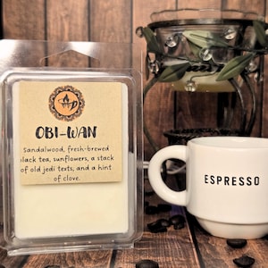 May include: A clear glass jar candle with a white wax base and a brown label that reads "OBI-WAN Sandalwood, fresh-brewed black tea, sunflowers, a stack of old jedi texts, and a hint of clove."