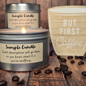May include: Two silver candle jars with brown paper labels that read "Sample Candle" and the text "Scent description will go here, so you know what it is you're sniffing." The candle on the left is lit. A wooden sign with the text "BUT FIRST Coffee" is in the background. Coffee beans are scattered on the wooden surface.