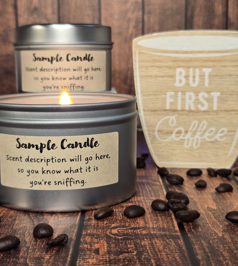 May include: Two silver candle jars with brown paper labels that read "Sample Candle" and the text "Scent description will go here, so you know what it is you're sniffing." The candle on the left is lit. A wooden sign with the text "BUT FIRST Coffee" is in the background. Coffee beans are scattered on the wooden surface.