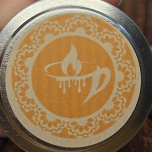May include: A silver metal lid with a beige label featuring a stylized orange flame design inside a circle with a decorative border. The label has the text "The Candle Bar" printed on it.
