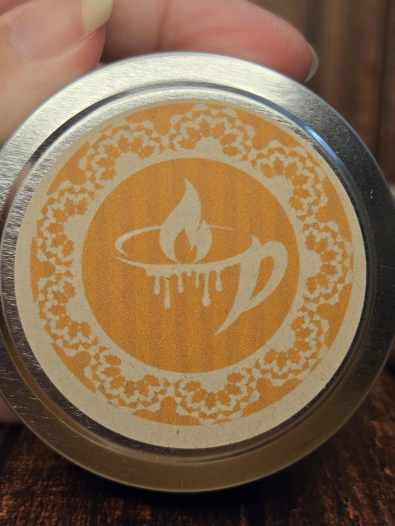 May include: A silver metal lid with a beige label featuring a stylized orange flame design inside a circle with a decorative border. The label has the text "The Candle Bar" printed on it.