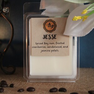May include: A white wax melt in a clear plastic container with a brown label that reads "Jesse Spiced Bay rum, frosted cranberries, sandalwood, and jasmine petals."
