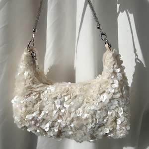 May include: A cream-coloured shoulder bag covered in shimmering white sequins. The bag features a top zip closure and a detachable strap encrusted with small, sparkling silver-toned crystals, attached to the bag with silver-toned metal clasps.