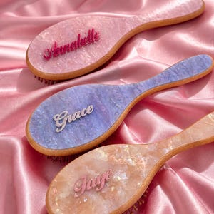 Puede incluir: Tres cepillos para el cabello personalizados con los nombres Annabelle, Grace y Faye. Cada cepillo presenta una combinación de colores única: rosa, azul y melocotón, con nombres a juego. Los cepillos tienen mangos de madera y están sobre un fondo de satén rosa.