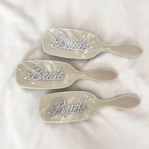 Puede incluir: Tres cepillos para el cabello de color beige claro con acabado marmolado, cada uno con la palabra Bride escrita con pequeños diamantes de imitación transparentes y reflectantes en la parte posterior del cabezal del cepillo.