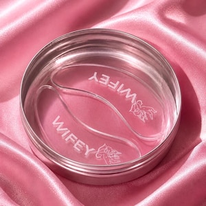 May include: A silver metal container holding two clear, curved gel pads. The pads are printed with the word "WIFEY" in white, and a small angel illustration. The background is a pink satin fabric.