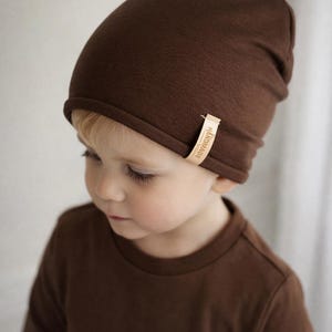 Könnte beinhalten: Ein brauner Beanie-Hut mit einem kleinen "HANDMADE"-Etikett wird von einem kleinen Kind getragen. Das Kind trägt auch ein braunes Langarmshirt. Der Hut und das Shirt sind aus einem weichen, einfarbigen Stoff.