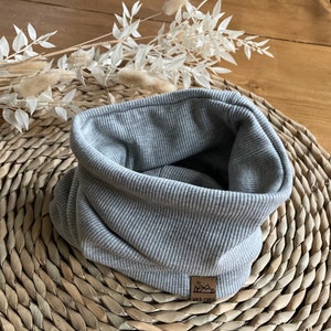 Könnte beinhalten: Ein grauer Rippstrick-Rundschal mit einem kleinen braunen Etikett mit der Aufschrift "WILD CHILD". Der Halswärmer ist aus einem weichen, dicken Material gefertigt und ist gefaltet. Der Hintergrund ist eine gewebte Matte.
