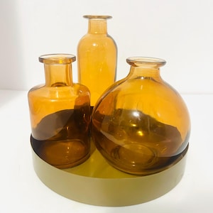 May include: Three amber glass vases of varying shapes sit on a gold-colored, circular tray. The vases are transparent, allowing light to pass through, and are arranged in a visually appealing composition. The background is plain white.