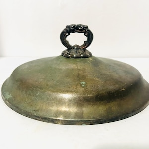 May include: A tarnished silver-toned metal serving dish lid. The lid is round with a decorative handle. The metal has a patina with green and brown hues. The handle is ornate with a dark finish.