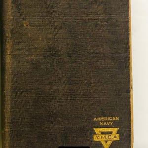 May include: A vintage copy of "The New Testament" with gold lettering on a dark brown cover. The cover features the text "AMERICAN NAVY" and the YMCA logo. The book is slightly worn, suggesting age.