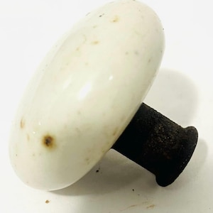 May include: A vintage-style door knob featuring a smooth, off-white ceramic handle with small brown speckles and signs of wear. The handle is attached to a dark, textured, cylindrical metal base that shows significant oxidation and rust.