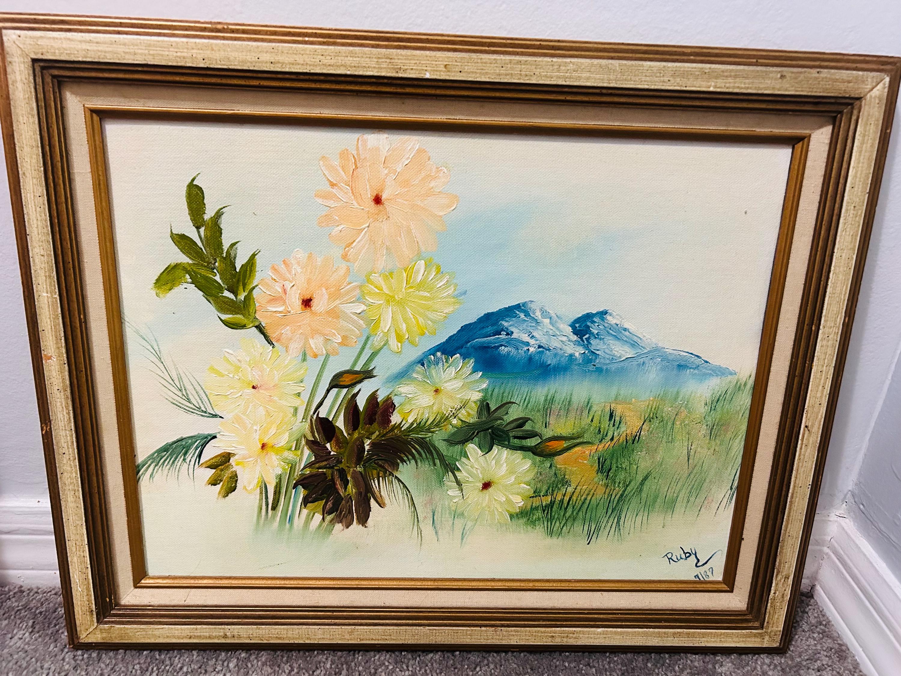 May include: An oil painting in a gold-toned frame depicts a landscape with flowers and mountains. The flowers are in shades of yellow and peach, with green leaves. Blue mountains are in the background, and the painting is signed "Ruby" with the year "9/87".