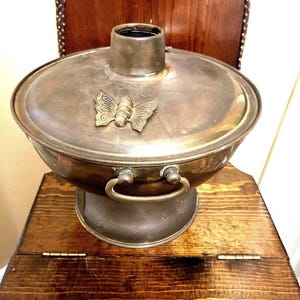 May include: A vintage brass hot pot with a butterfly design on the lid. The pot has two handles and sits on a wooden stand.