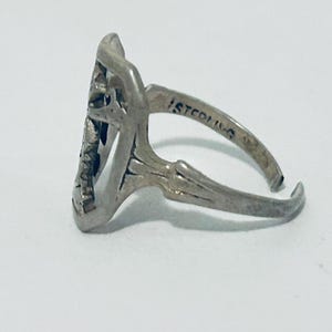 May include: A silver-colored ring with an openwork design featuring a central motif. The band is stamped with the word STERLING on the inner surface. The ring has an adjustable open shank construction with a metallic finish and visible patina.