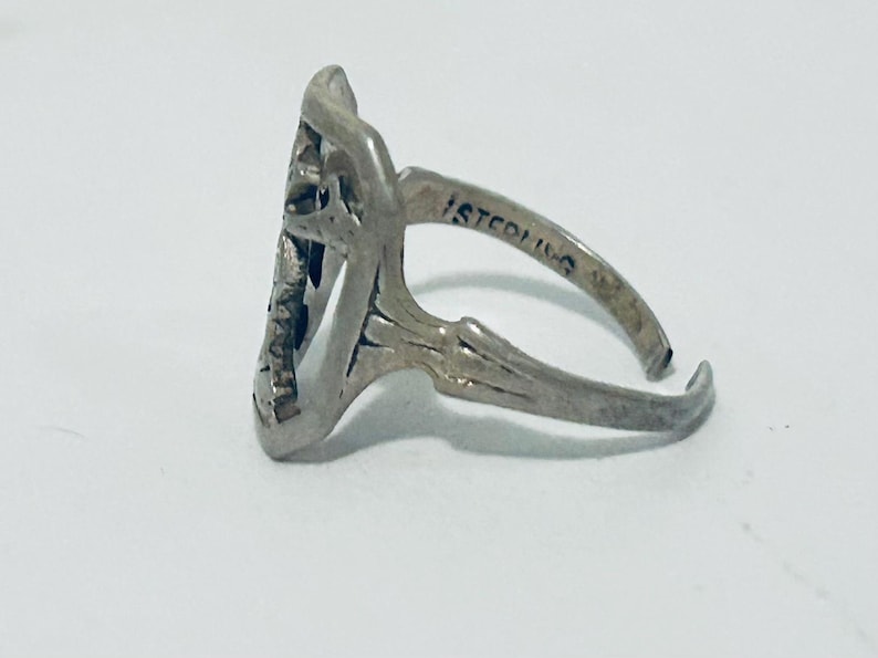 May include: A silver-colored ring with an openwork design featuring a central motif. The band is stamped with the word STERLING on the inner surface. The ring has an adjustable open shank construction with a metallic finish and visible patina.