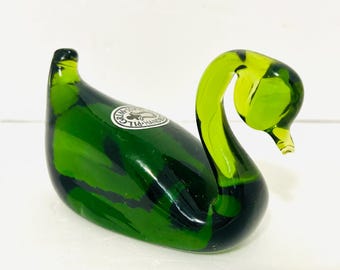 May include: A handcrafted glass swan figurine in shades of green. The swan's body is a deep, forest green, while the neck and head are a lighter, lime green. A circular sticker is visible on the body. The swan is a decorative item.