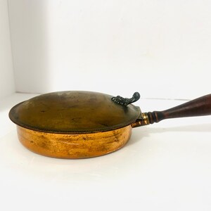 May include: Antique copper pan with a wooden handle. The pan has a lid with a decorative metal handle. The copper has a warm, aged patina. The handle is dark brown and attached with brass hardware. A vintage kitchenware item.