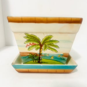 May include: A square ceramic planter with a matching saucer featuring a hand-painted tropical scene. The design includes a green palm tree with red fruit, a blue ocean, yellow sailboats, and a brown border along the top and bottom edges.
