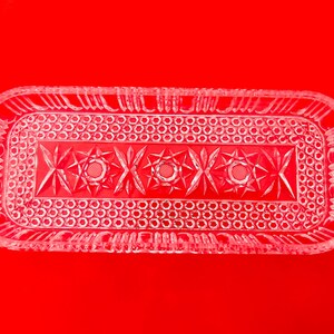 May include: A rectangular clear glass tray featuring a pressed pattern with a central row of starburst motifs surrounded by a border of small circular indentations and vertical fluted edges along the outer rim of the dish.