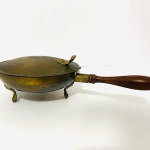 May include: Antique brass warming pan with a wooden handle. The pan has a rounded shape with a lid and three small feet. The handle is dark brown and the pan has a tarnished, aged appearance.