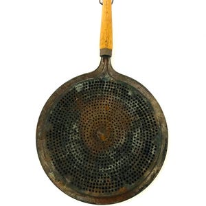 May include: An antique, round metal kitchen tool with a wooden handle. The tool has a perforated surface with numerous small holes. The handle is light brown, and the metal has a rustic, aged appearance. A metal loop is attached to the handle.