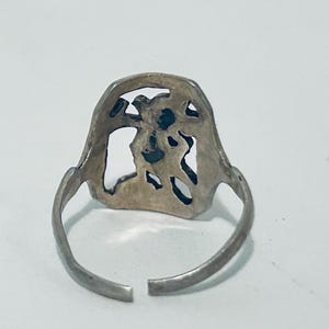 May include: A silver-toned metal ring featuring an openwork design on the face. The ring has a thin, adjustable band with a gap at the bottom, showcasing a cutout pattern that resembles a stylized figure or abstract shape within the frame.