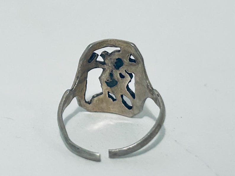 May include: A silver-toned metal ring featuring an openwork design on the face. The ring has a thin, adjustable band with a gap at the bottom, showcasing a cutout pattern that resembles a stylized figure or abstract shape within the frame.