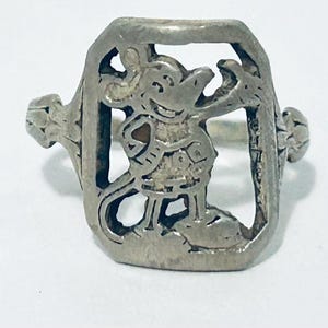 May include: A silver-toned metal ring featuring a rectangular frame with a cutout design of a cartoon mouse character. The mouse is depicted in a standing position with one arm raised, enclosed within the metallic border of the ring's face.