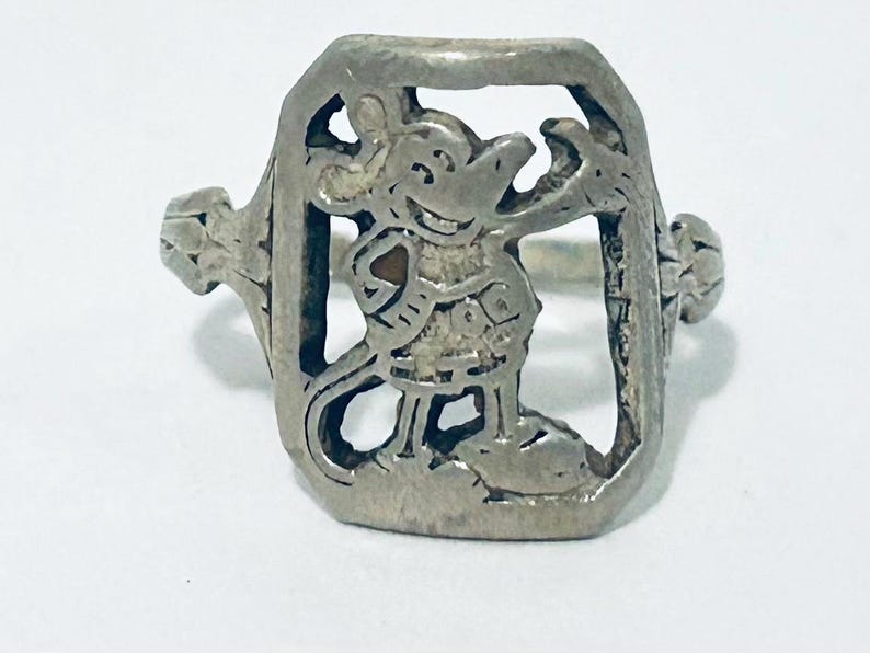 May include: A silver-toned metal ring featuring a rectangular frame with a cutout design of a cartoon mouse character. The mouse is depicted in a standing position with one arm raised, enclosed within the metallic border of the ring's face.