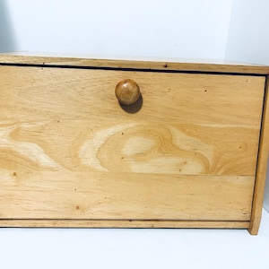 May include: A rectangular light brown wooden bread box with a smooth finish and a visible natural wood grain pattern. The front panel features a single centered round wooden knob used for opening the hinged door of the storage container.