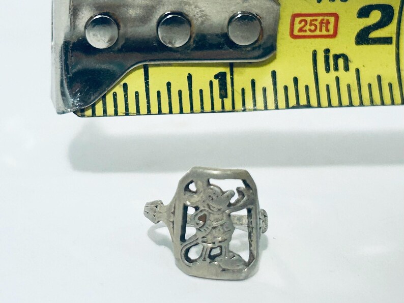 May include: A silver-toned metal ring featuring a cutout design of a cartoon mouse character. The ring is positioned below a yellow measuring tape, which indicates the ring's height is approximately 0.5 inches from the top to the bottom edge.