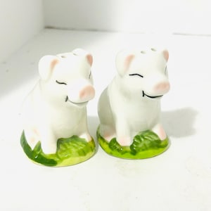 May include: Two ceramic pig salt and pepper shakers. The pigs are white with pink ears, snouts, and feet. They sit on a green and yellow base, with a smiling face and closed eyes.