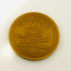 May include: A gold-colored commemorative coin with the text "Centennial" at the top. The coin features an image of a bison and the words "Nebraskaland" and "where the West begins 1867-1967".