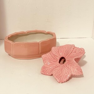 May include: A pink ceramic trinket box shaped like a flower with a matching lid. The lid is a stylized flower with a hole in the center. The box is oval shaped with a scalloped edge.