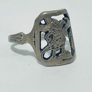May include: A silver-toned metal ring featuring a rectangular face with a cut-out design of a cartoon mouse character. The mouse is depicted in a standing position with its hand near its head. The band of the ring has a simple, tapered design.