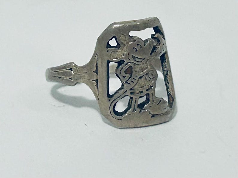 May include: A silver-toned metal ring featuring a rectangular face with a cut-out design of a cartoon mouse character. The mouse is depicted in a standing position with its hand near its head. The band of the ring has a simple, tapered design.